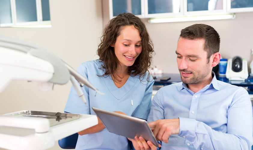 Dental nurse and male patient reviewing treatment plan on a digital tablet in a modern surgery clinic.