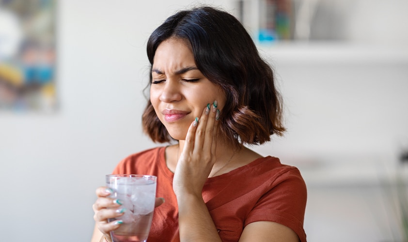 Woman wincing in pain from sensitive teeth while holding a glass of ice water, illustrating dental hypersensitivity symptoms.
