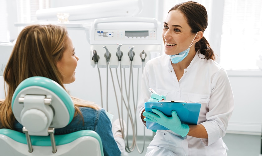 Smiling dentist with clipboard talking to a patient in a dental chair, providing professional consultation in a modern clinic.