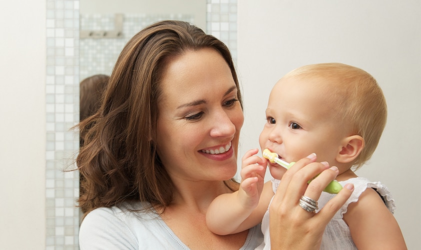 Mother smiling at toddler using a colorful toothbrush for infant dental hygiene and oral care.