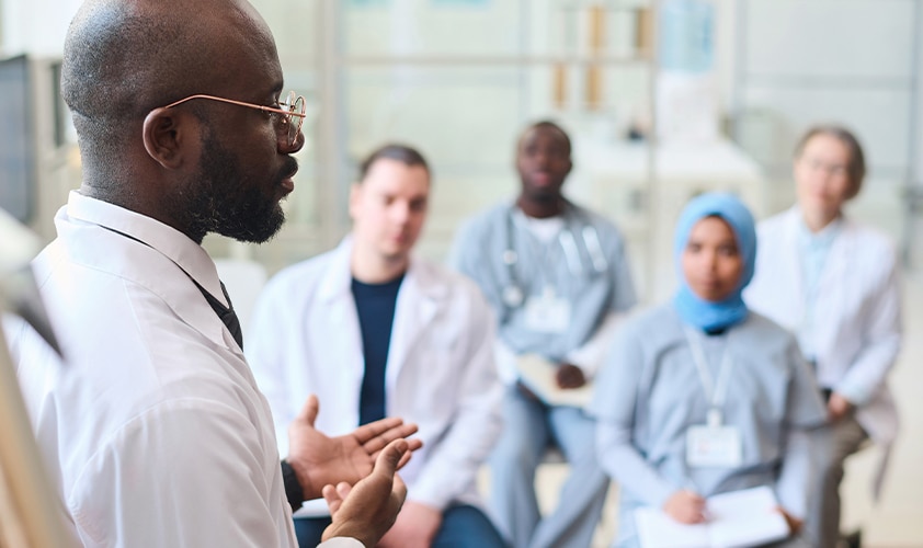 Doctor leading a medical seminar for a diverse group of healthcare professionals and students in a clinical training environment.
