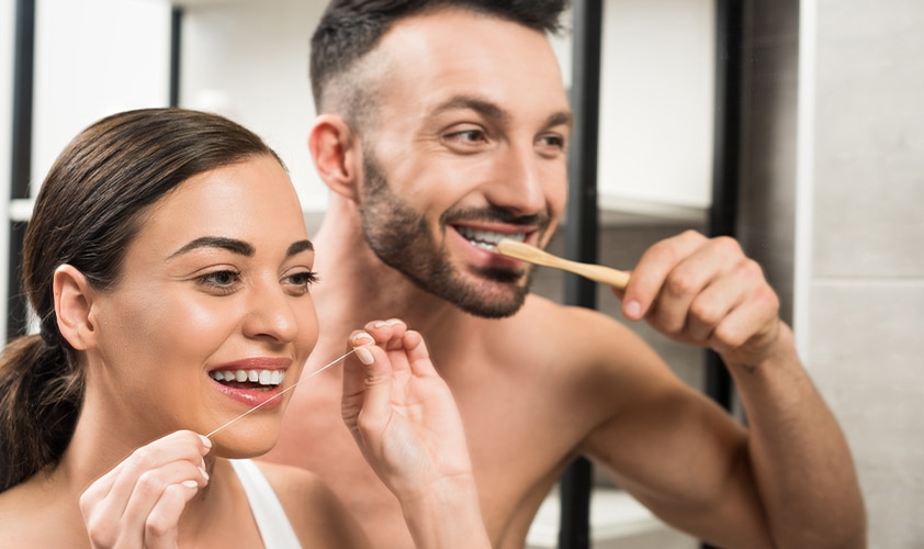 Smiling couple performing daily oral hygiene; woman flossing and man brushing teeth with a bamboo toothbrush in front of a bathroom mirror.