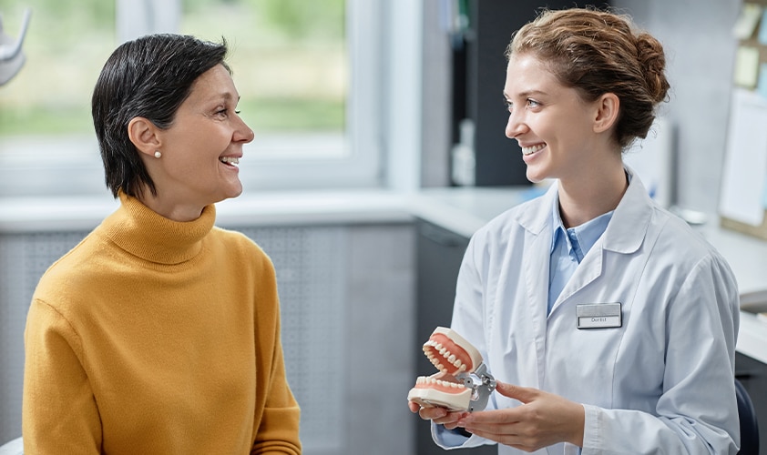 Dentist holding a denture model while explaining a dental procedure to a smiling patient during a consultation at a dental clinic.
