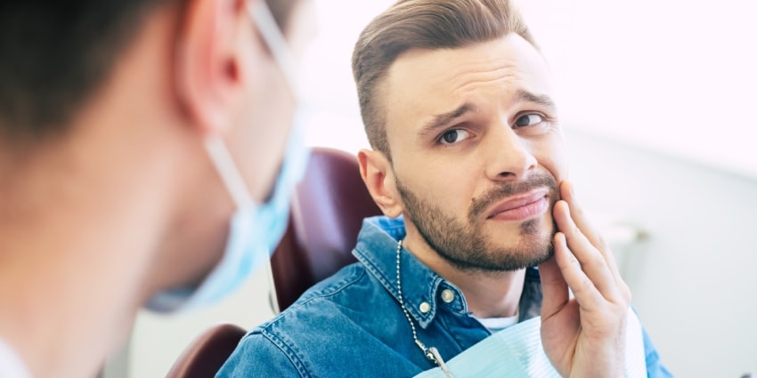 A man with a worried face is holding his hand on his cheek  because of irritating pain in front of a dentist who is going to give a patient a treatment.